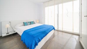 Ein Schlafzimmer mit einem Bett, einem Nachttisch, einer Lampe und einem großen Fenster mit Vorhängen.