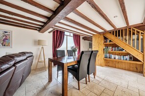 A dining area with a wooden table and chairs, a leather sofa, a lamp, and a bookshelf with books and decorative items. - Granary Barn, A Rural Retreat - Private Hot Tub, (Winkleigh)