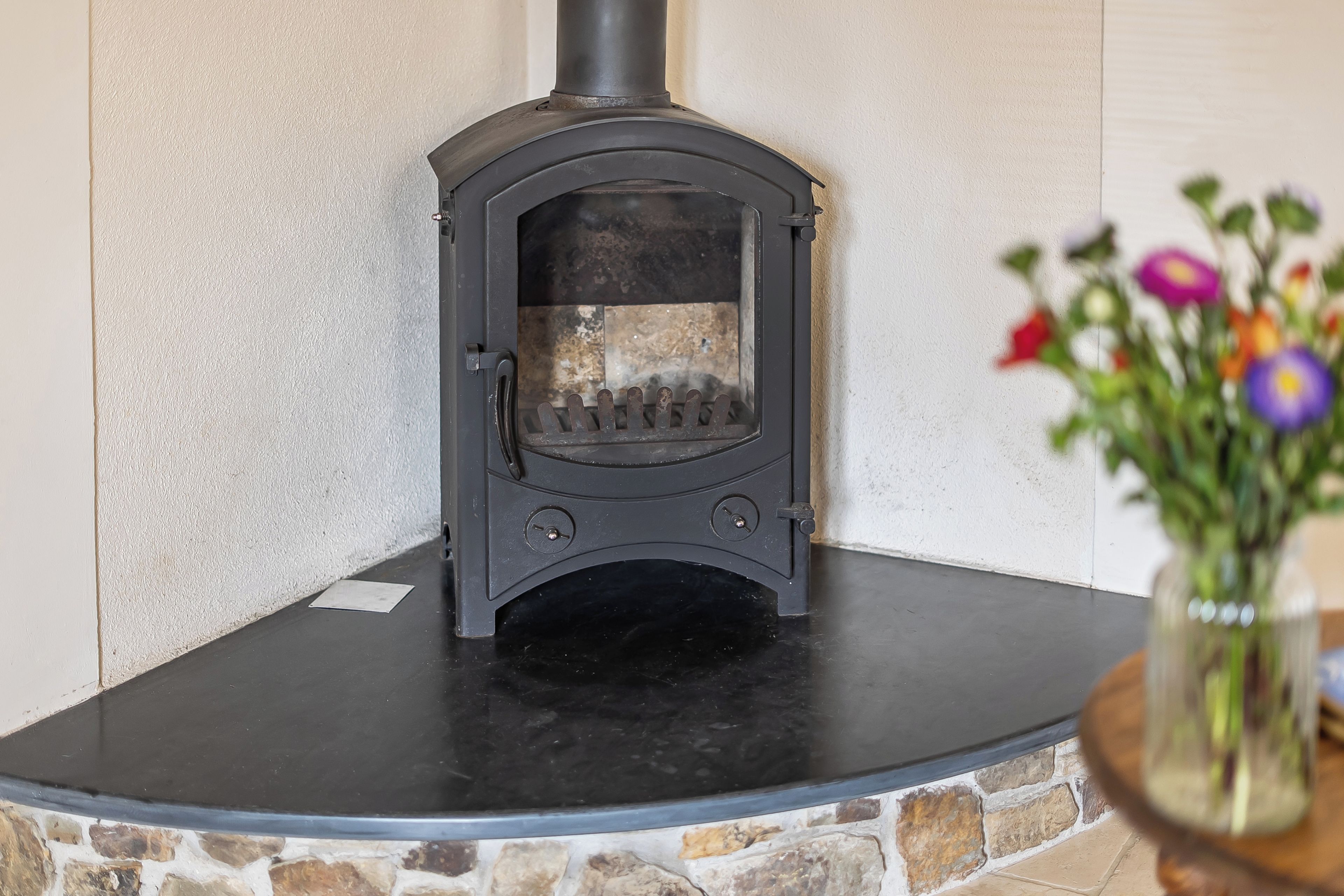 A wood-burning stove with a stone surround, a small table with a vase of flowers, and a white textured wall.