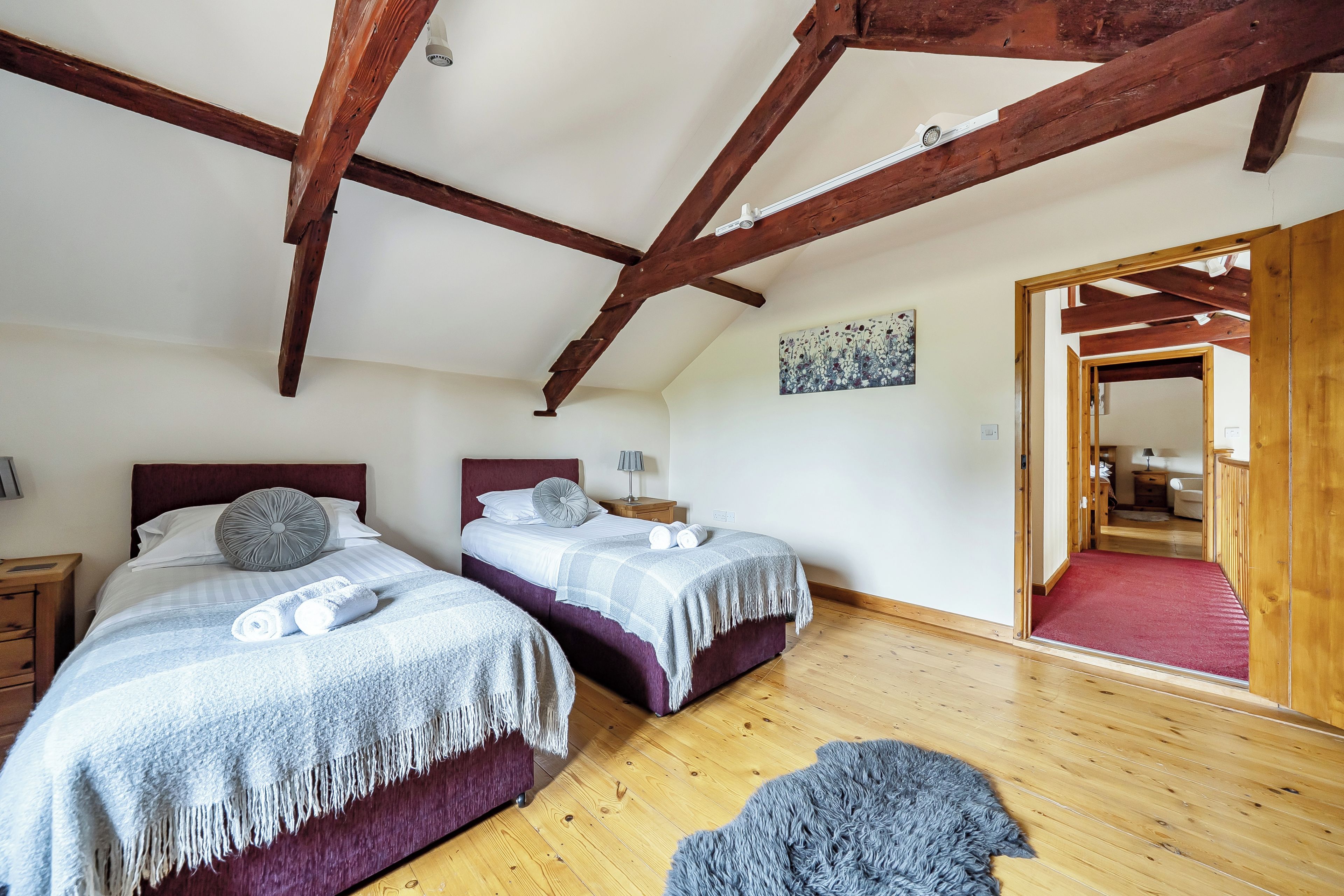 A bedroom with two beds, wooden beams on the ceiling, a painting on the wall, and a hallway leading to another room.