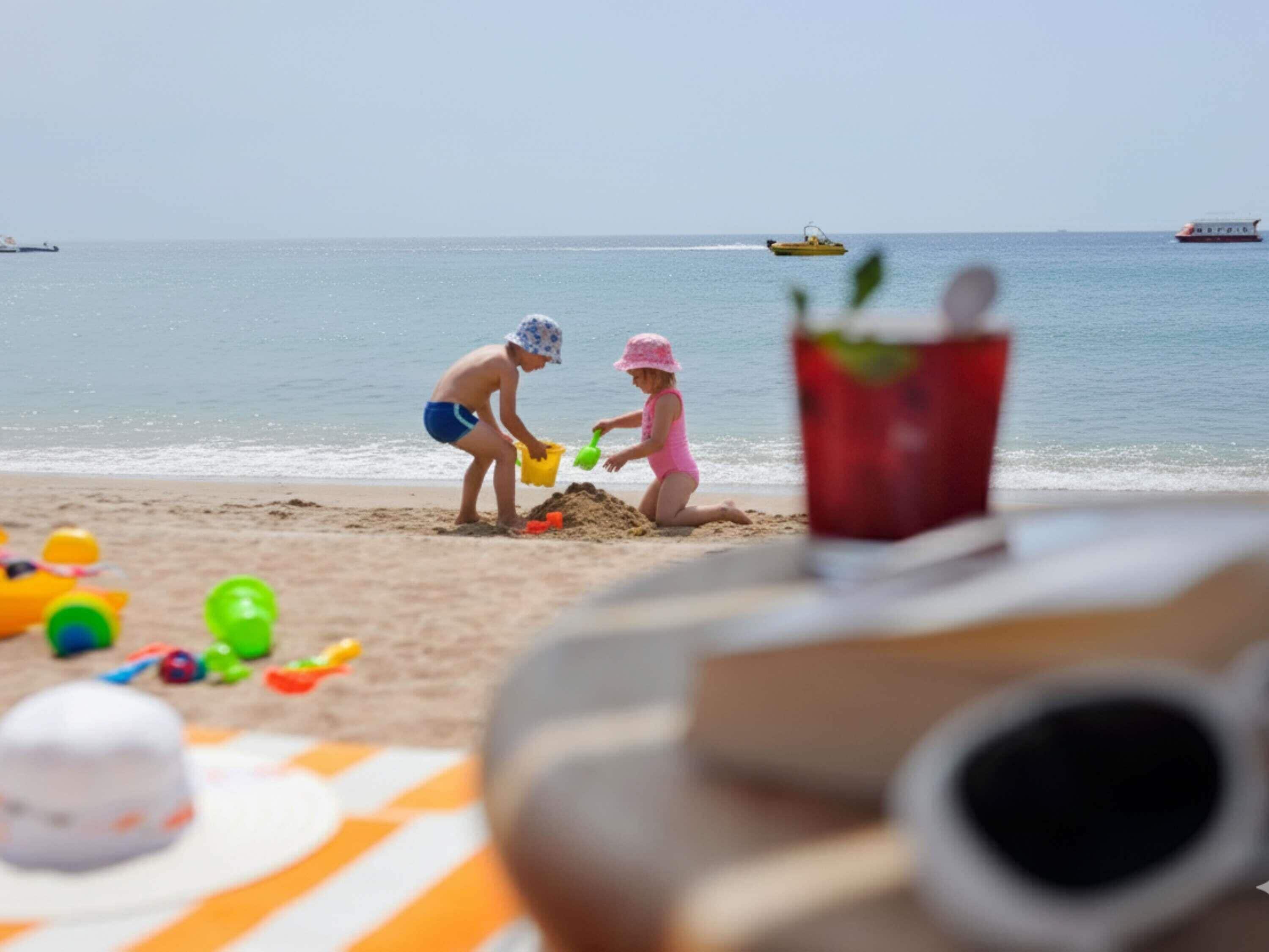 A beach scene with two children playing in the sand, a red cup, and beach toys.
