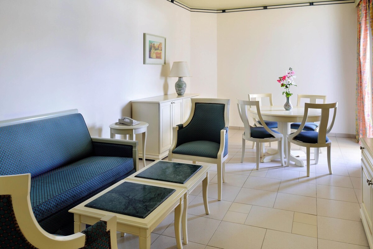 A living room with a sofa, two armchairs, a coffee table, a dining table with chairs, a lamp, and a painting on the wall.