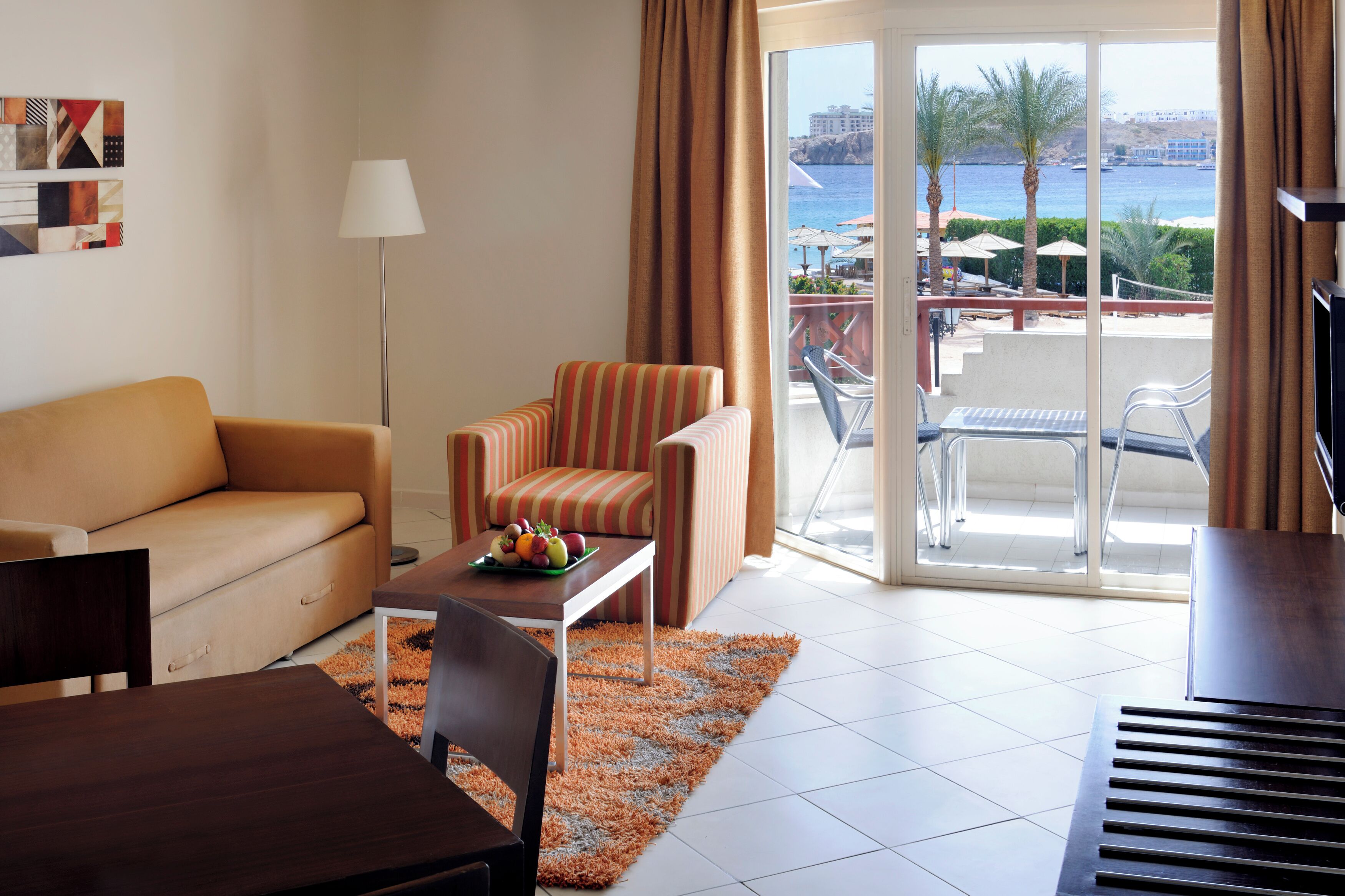 Une chambre d’hôtel avec un canapé, un fauteuil à rayures, une table à manger et une vue sur la mer, visible à travers une porte-fenêtre coulissante.
