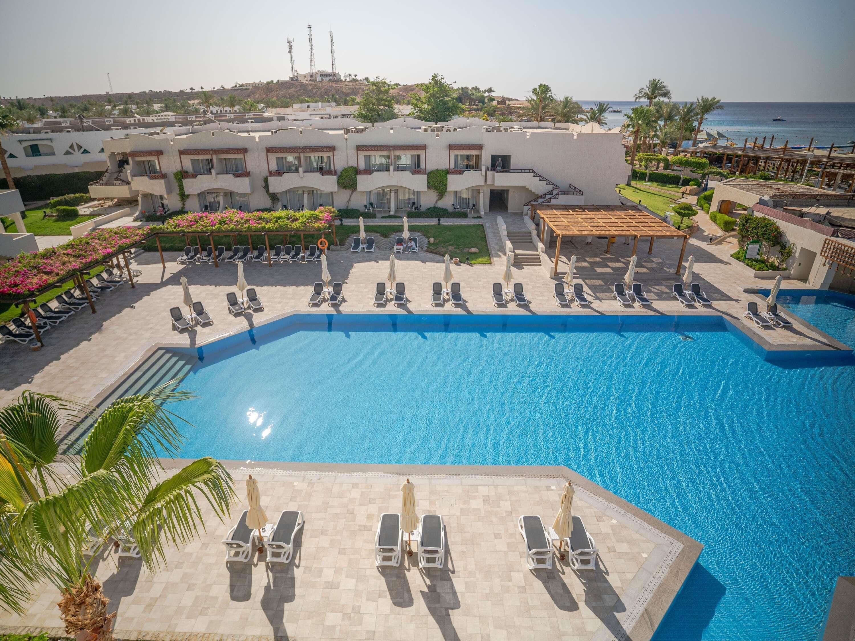 A large swimming pool with sun loungers and umbrellas, surrounded by a building with multiple balconies and a view of the sea.