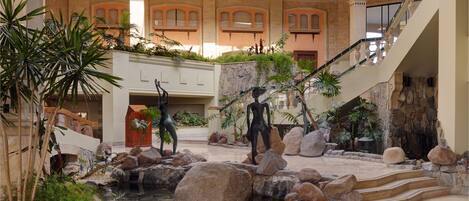 A spacious indoor atrium with a central water feature, stone walls, and a glass ceiling allowing natural light.