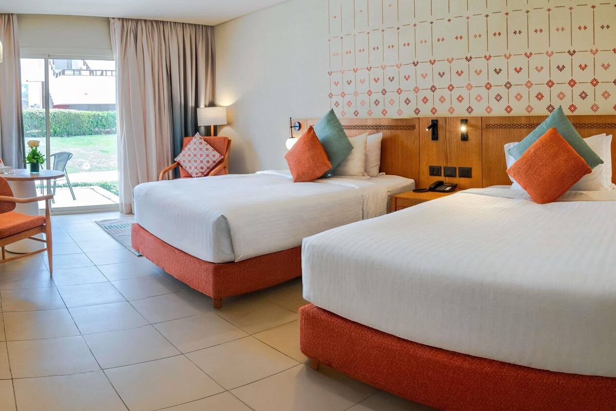 A hotel room with two beds, a sitting area with a chair and table, and a view of the outdoors through a large window.