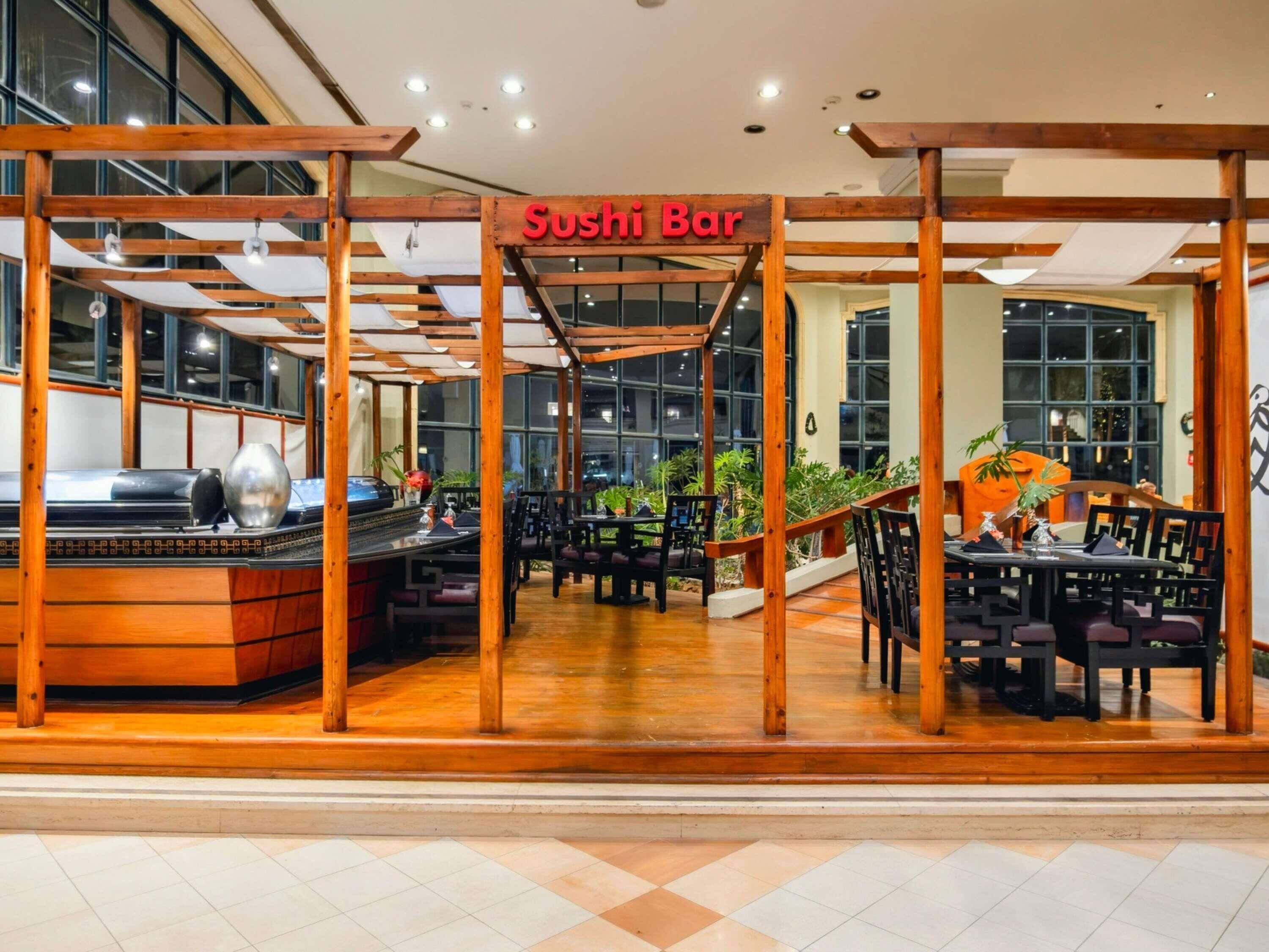 Un bar à sushis avec des poutres en bois et une enseigne, entouré de tables et de chaises, avec de grandes fenêtres en arrière-plan.