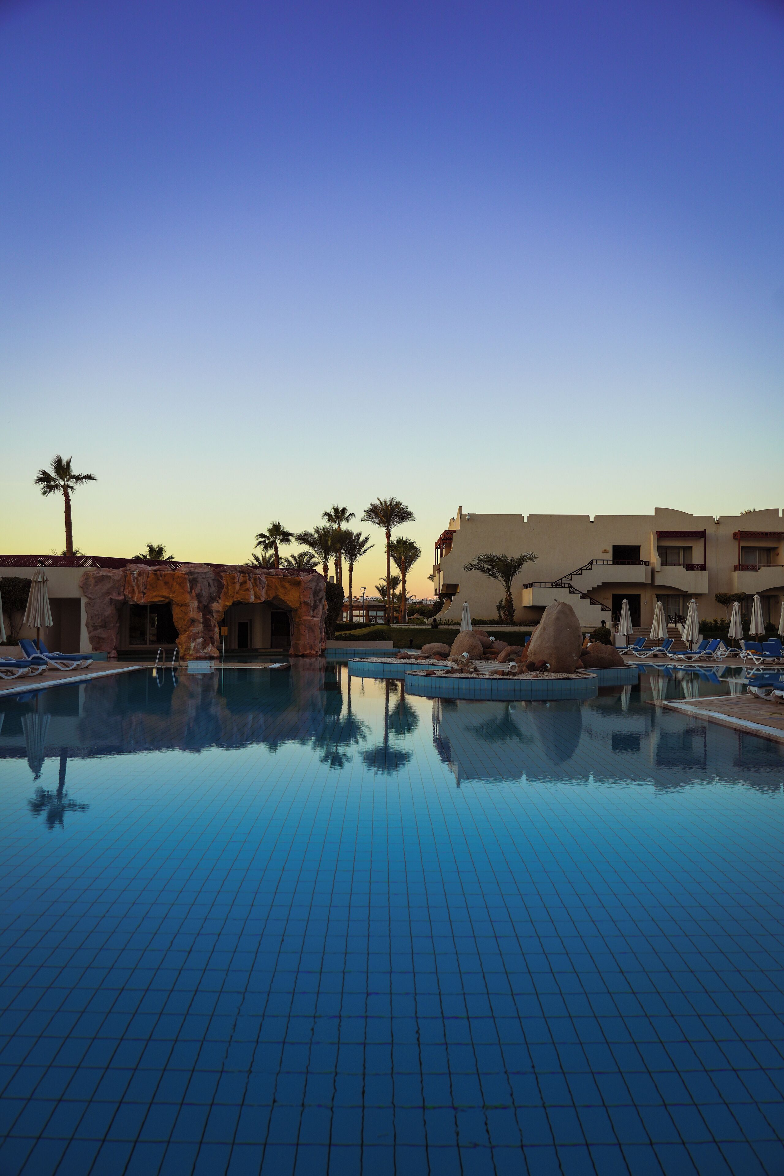 A hotel pool area with clear blue water, lounge chairs, palm trees, and a building with a rock facade.