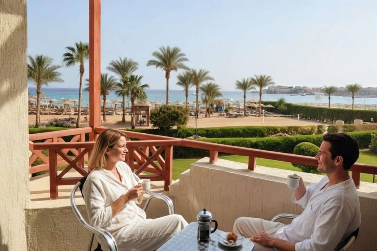 A couple seated at a table on a balcony overlooking a beach with palm trees and a clear sky.