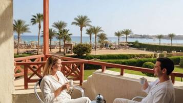 A couple seated at a table on a balcony overlooking a beach with palm trees and a clear sky.