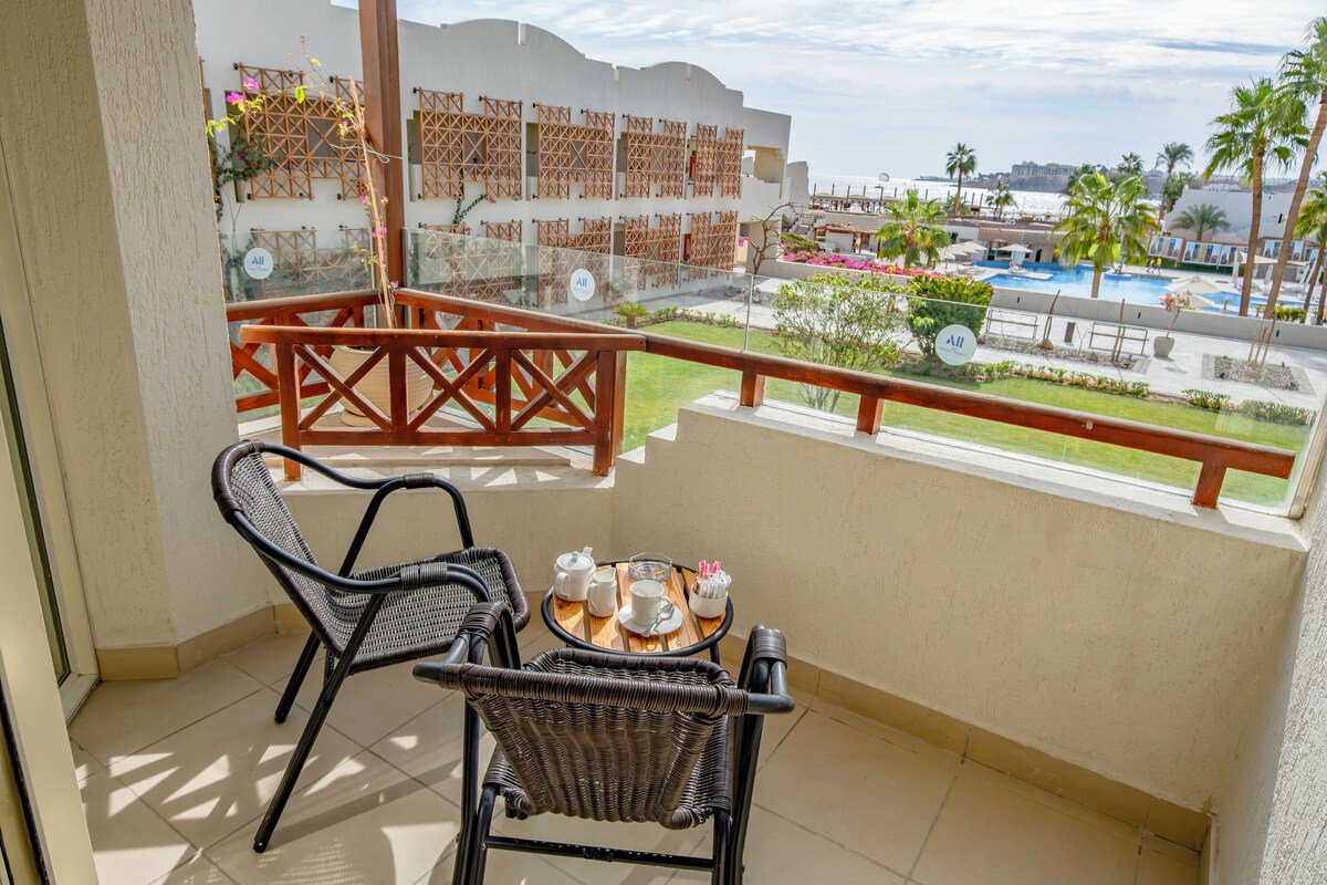 Un balcón con sillas de mimbre y una mesa, con vistas a una piscina y palmeras.