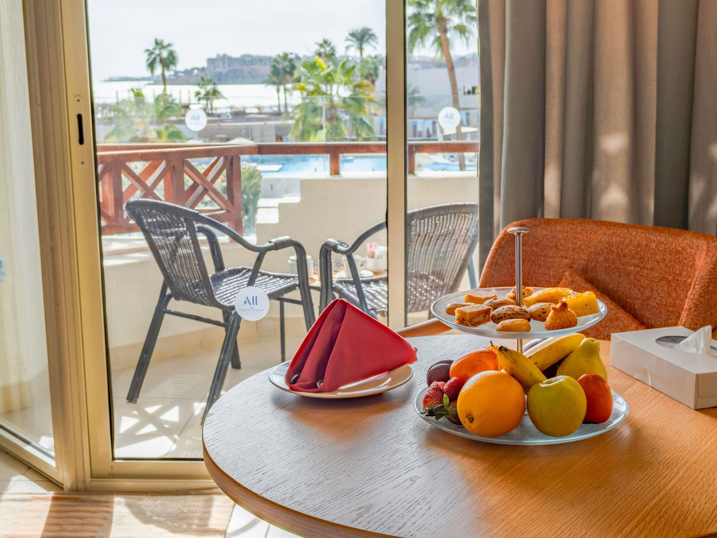 Ein Esstisch mit Obstteller, Servietten und Taschentuchbox auf einem Balkon mit Blick auf einen Pool und Palmen.