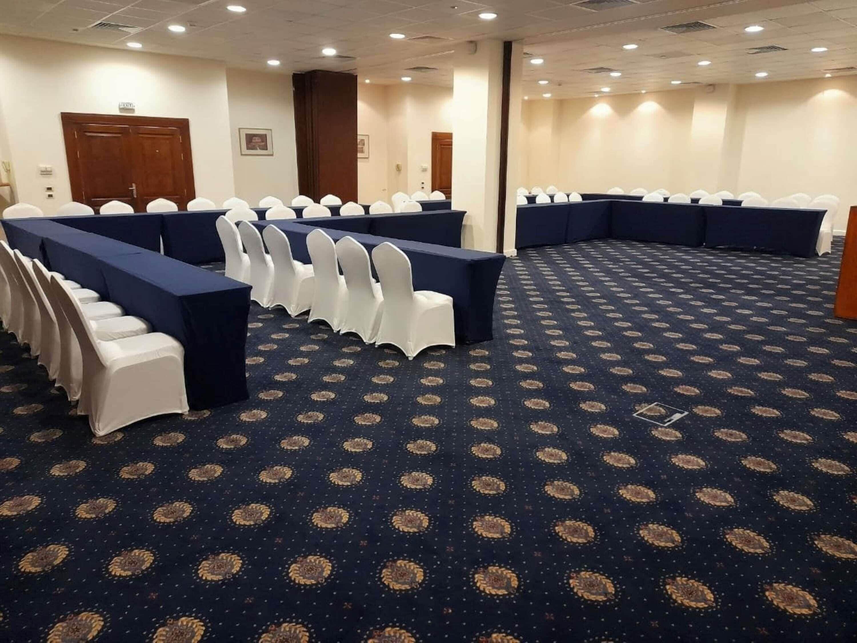 A conference room with rows of tables and chairs, a patterned carpet, and a door in the background.