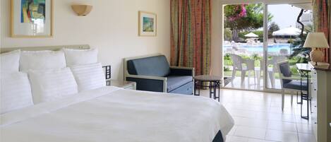 A hotel room with a large bed, a sofa, a small table, and a balcony with a view of a pool and greenery.