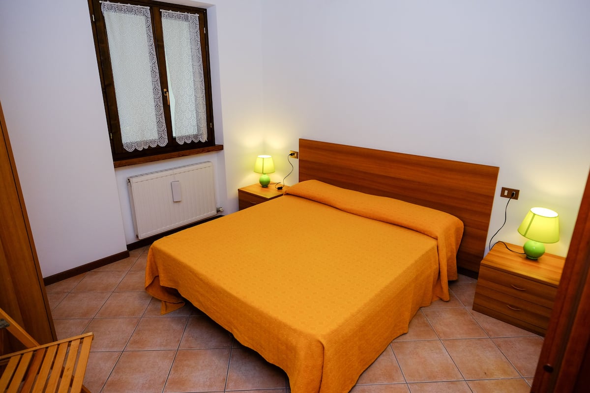 Une chambre à coucher avec un lit en bois, deux tables de chevet avec des lampes, une fenêtre avec des rideaux en dentelle et un radiateur.