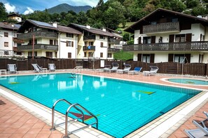 A swimming pool with tiled flooring and a metal ladder, surrounded by lounge chairs and a wooden fence. - Residence Alessio (Ledro)