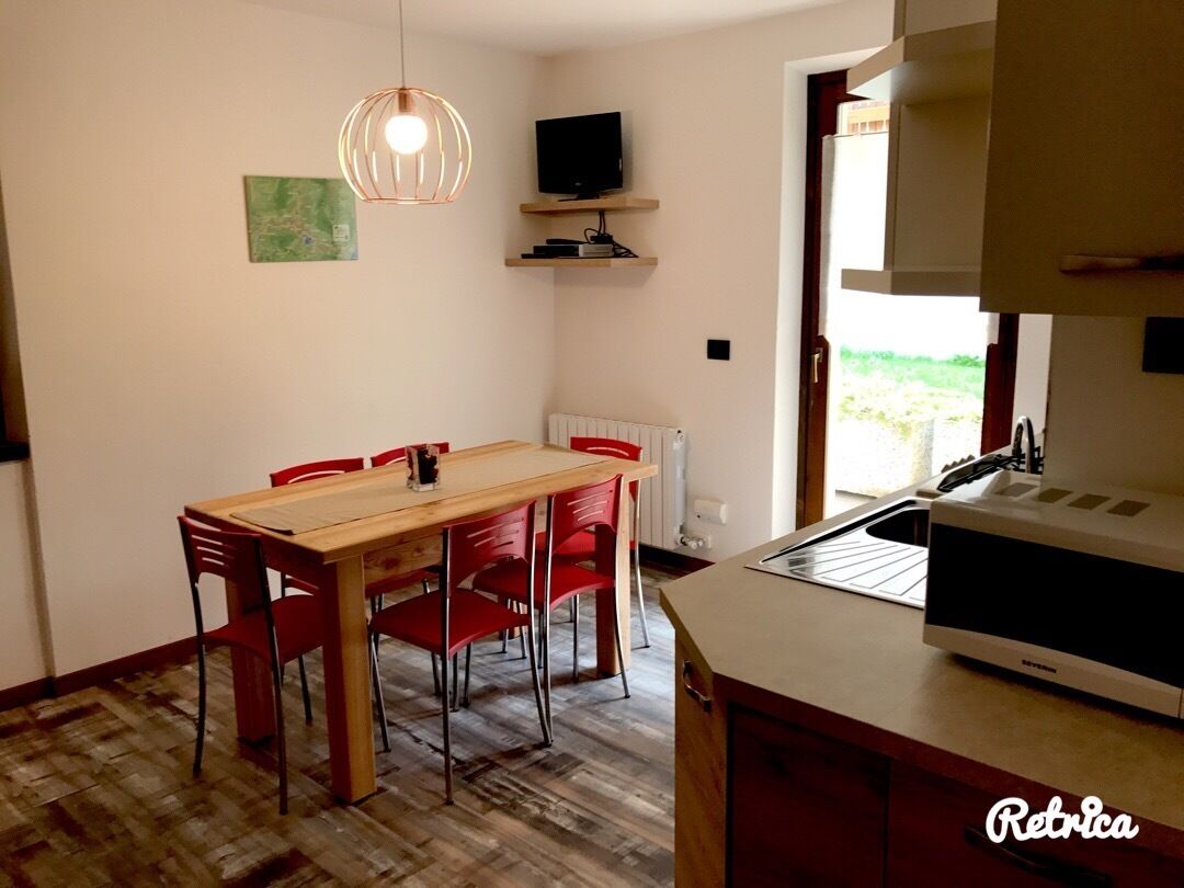 Un coin repas avec une table en bois et des chaises rouges, un luminaire suspendu, un téléviseur posé sur une étagère, et un espace cuisine équipé d’un four à micro-ondes.