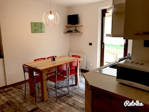A dining area with a wooden table and red chairs, a hanging light fixture, a television on a shelf, and a kitchen area with a microwave. - Residence Alessio (Ledro)