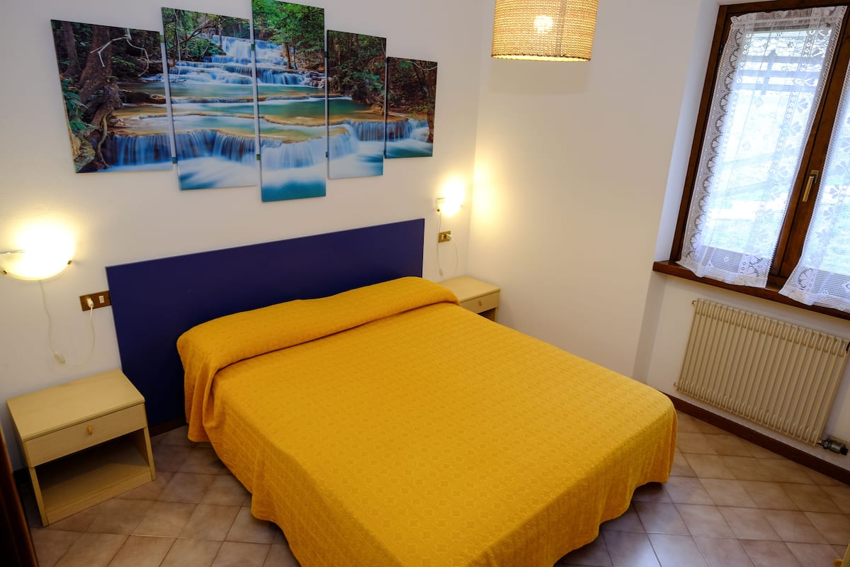 Une chambre à coucher comprenant un lit, des tables de chevet, une lampe fixée au mur, une fenêtre avec des rideaux en dentelle et un radiateur.