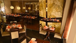 A restaurant with multiple tables set with orange napkins, white plates, and cutlery. The interior features dark wooden furniture, patterned wallpaper, and hanging pendant lights. - Hotel Regency Square (Gwalior)