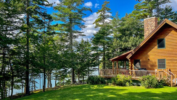 A log cabin with a deck and a stone chimney, surrounded by trees and a lake. - Waterfront log cabin nestled in the woods providing unparalleled privacy (Harpswell)