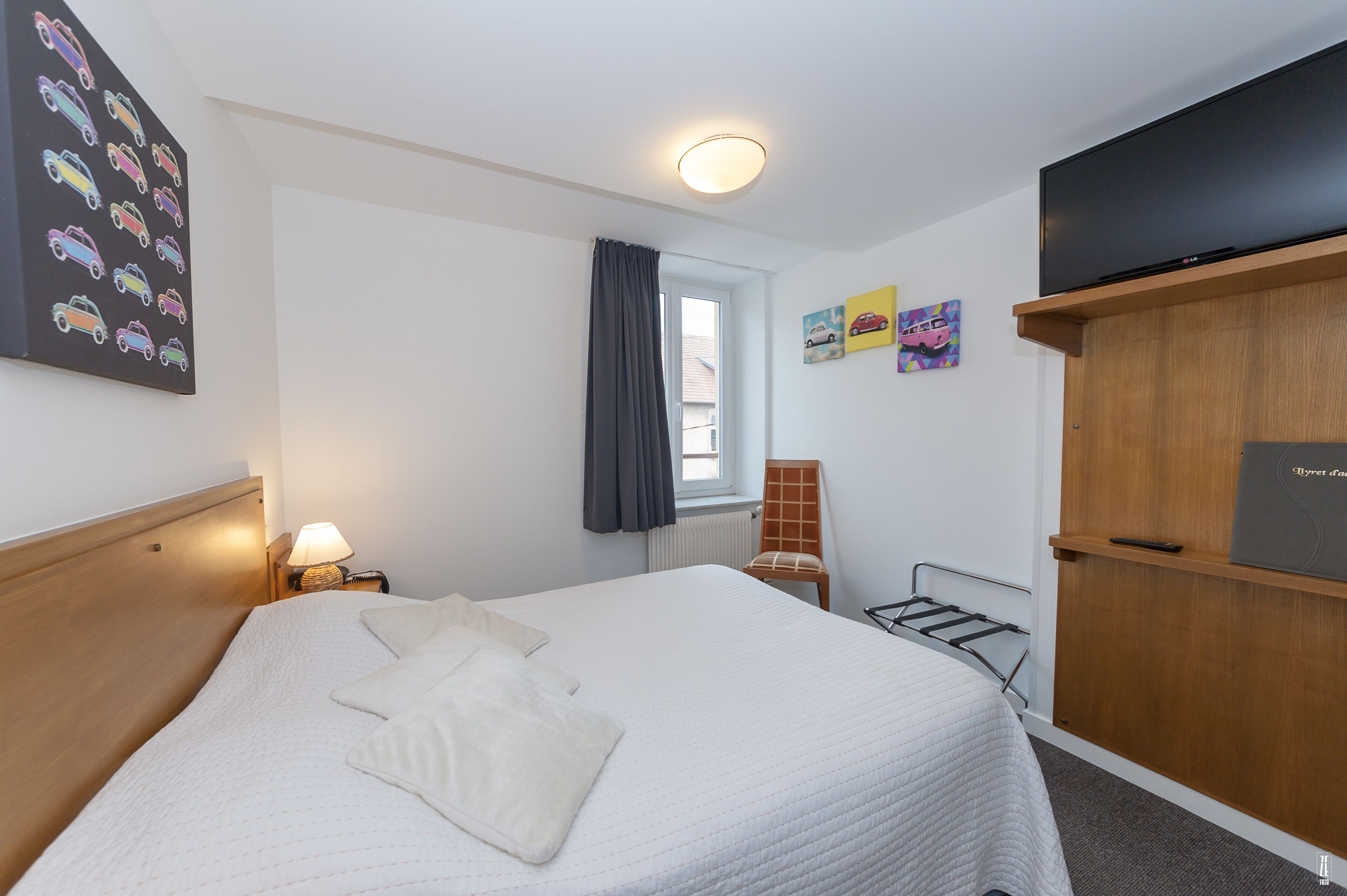 A neatly made bed with pillows, a bedside lamp, a window with curtains, a radiator, a chair, and a TV on a wooden cabinet.
