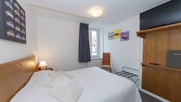 A neatly made bed with pillows, a bedside lamp, a window with curtains, a radiator, a chair, and a TV on a wooden cabinet.