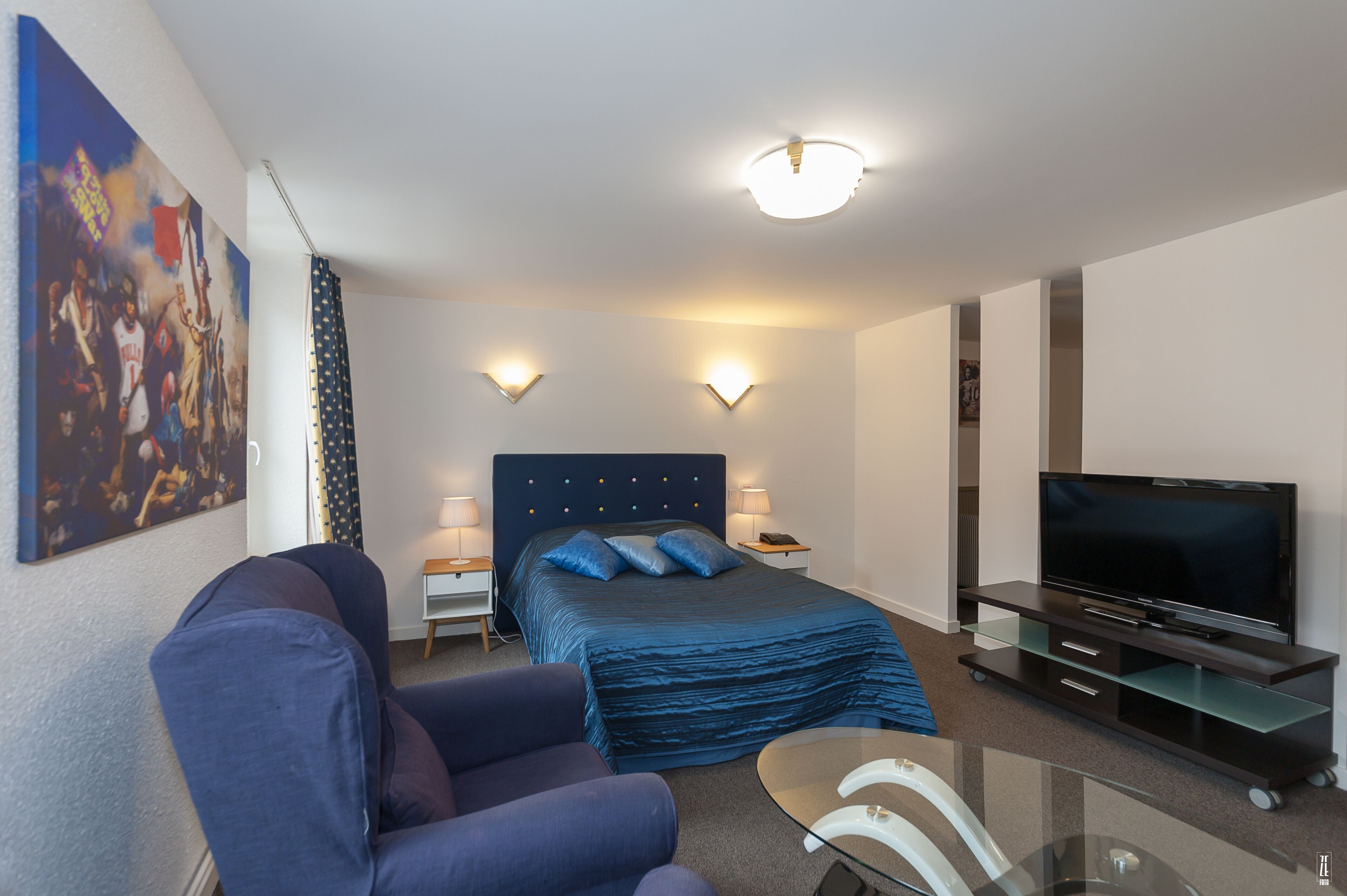 Una habitación de hotel con cama, televisión, sillón y un cuadro en la pared.