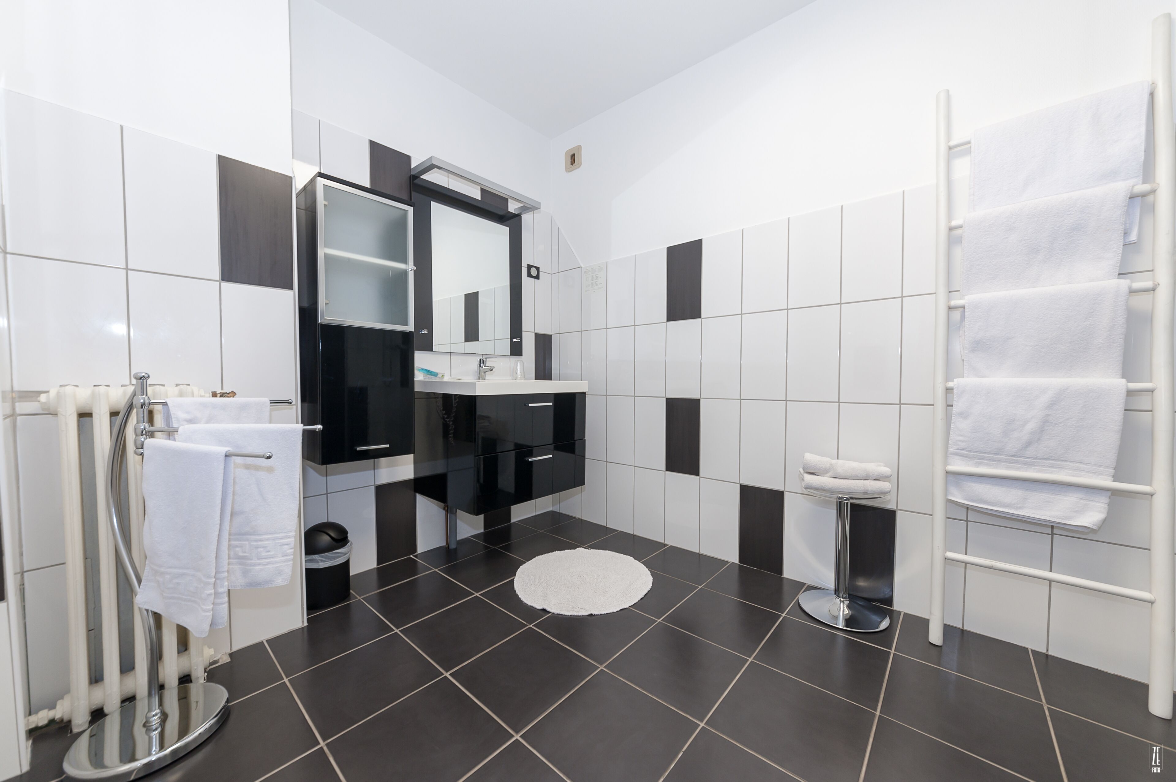 A modern bathroom with a black and white tiled floor, a round white rug, a white towel rack, a black vanity with a mirror, and a white towel rack.