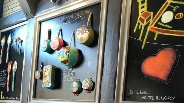 A restaurant interior with a dining table set for two, featuring a black napkin, a fork, and a glass. The wall behind the table displays colorful kitchen utensils and a chalkboard with French text. - Relais Lorraine Alsace (Raon-l'Etape)