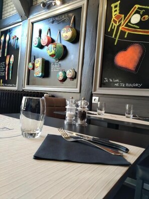 A restaurant interior with a dining table set for two, featuring a black napkin, a fork, and a glass. The wall behind the table displays colorful kitchen utensils and a chalkboard with French text. - Relais Lorraine Alsace (Raon-l'Etape)