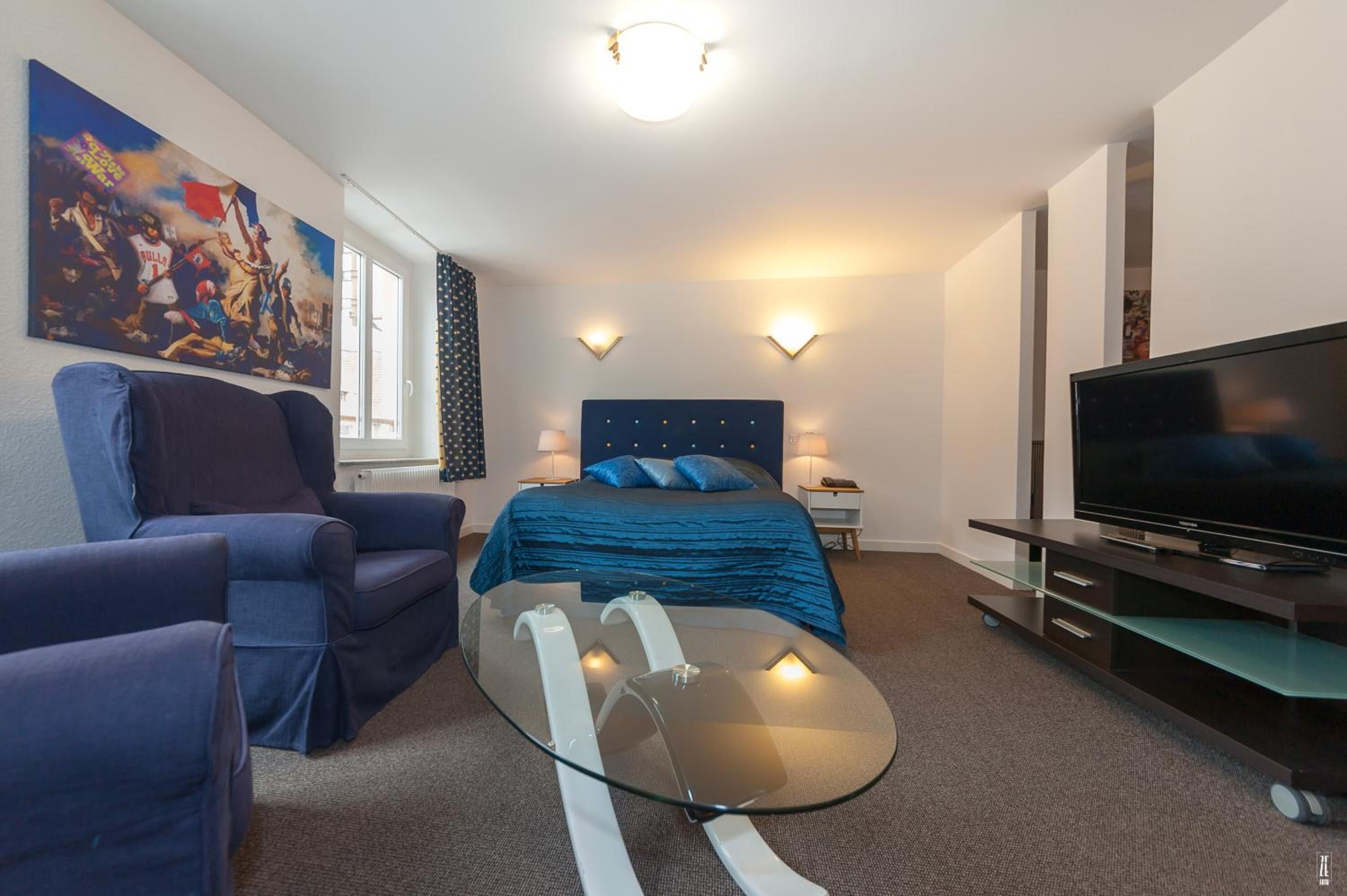 A modern hotel room with a bed, a glass coffee table, a TV, and a seating area with two armchairs.
