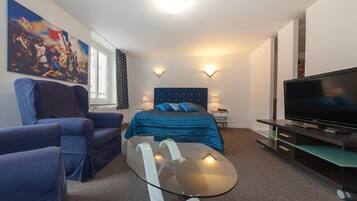 A modern hotel room with a bed, a glass coffee table, a TV, and a seating area with two armchairs.