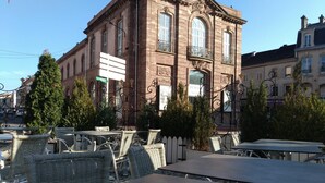 An outdoor cafe with wicker furniture and a building with arched windows in the background. - Relais Lorraine Alsace (Raon-l'Etape)