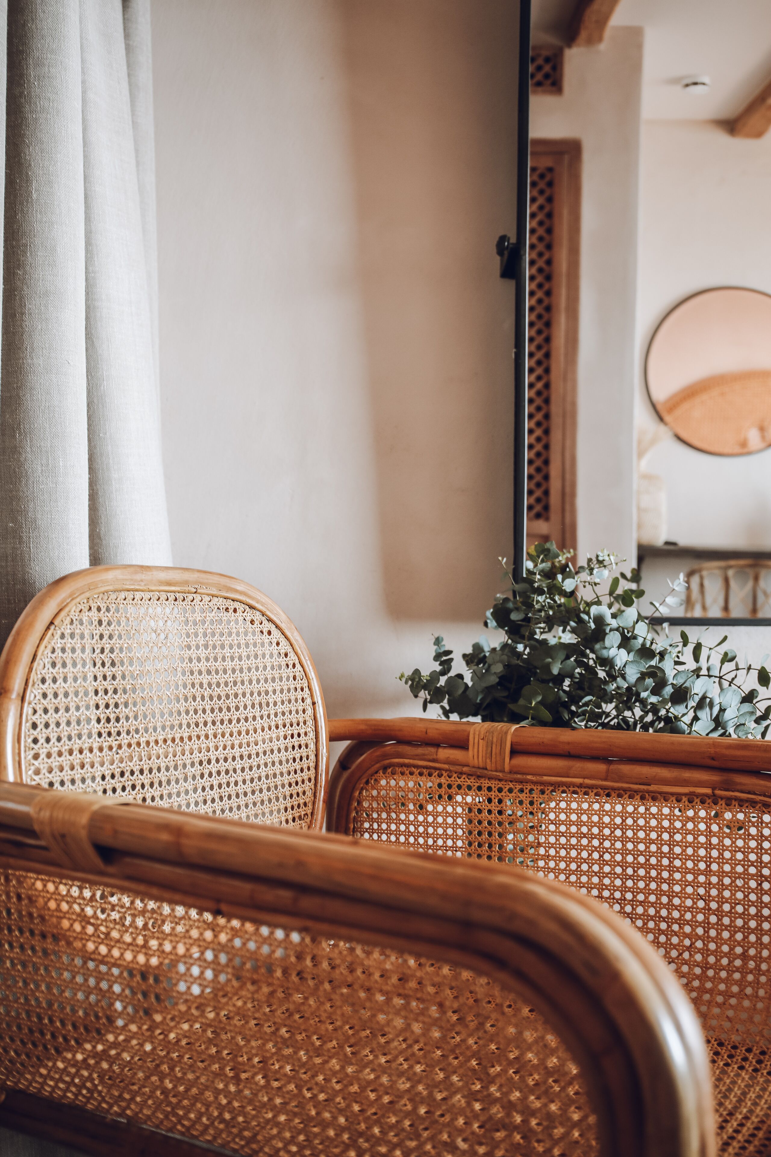 A wicker chair with a woven seat and backrest, positioned in front of a wall with a wooden door and a round mirror reflecting a plant.