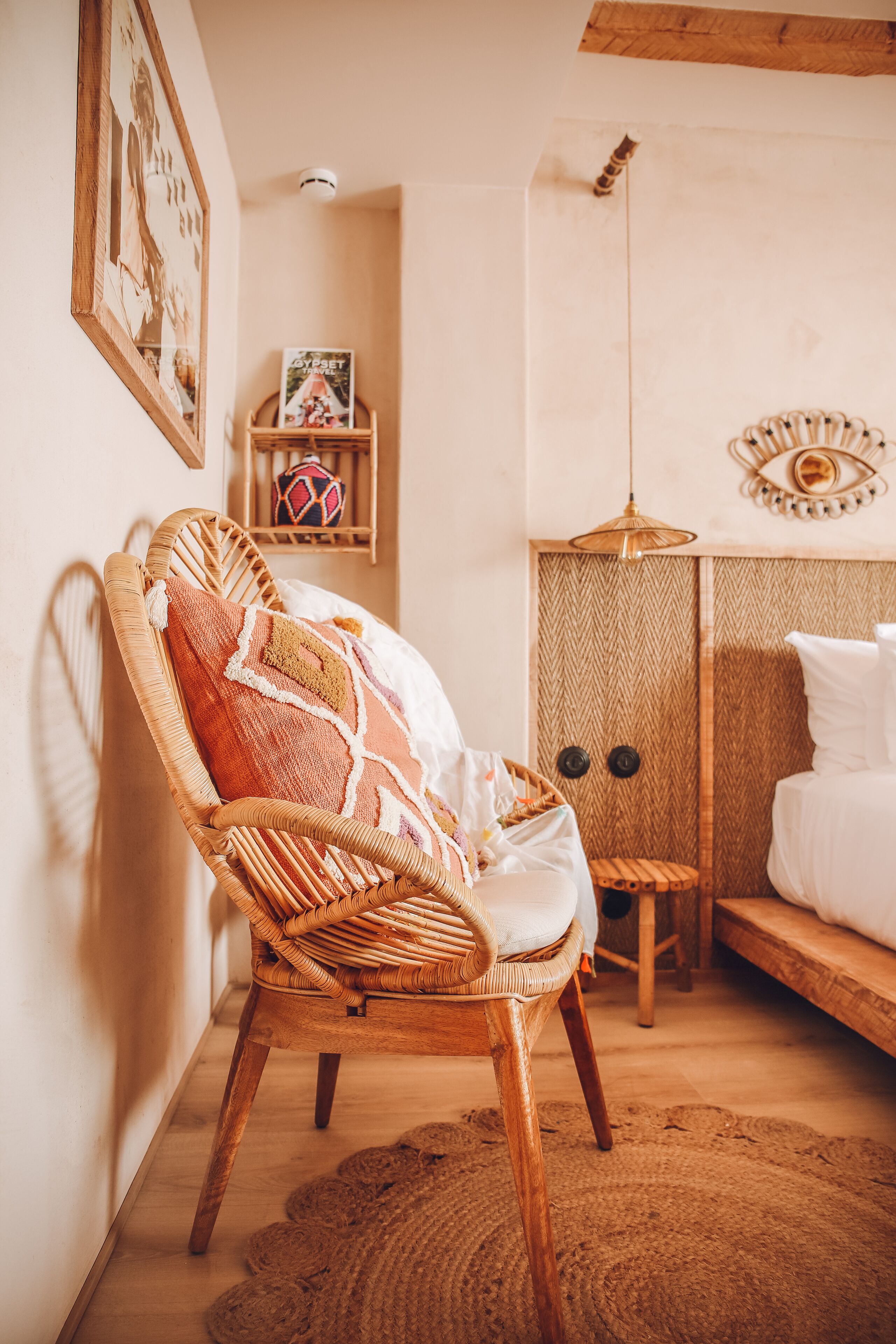 Una habitación acogedora con una silla de mimbre, una cama, un tapiz tejido, una estantería con un libro y un adorno decorativo en la pared.