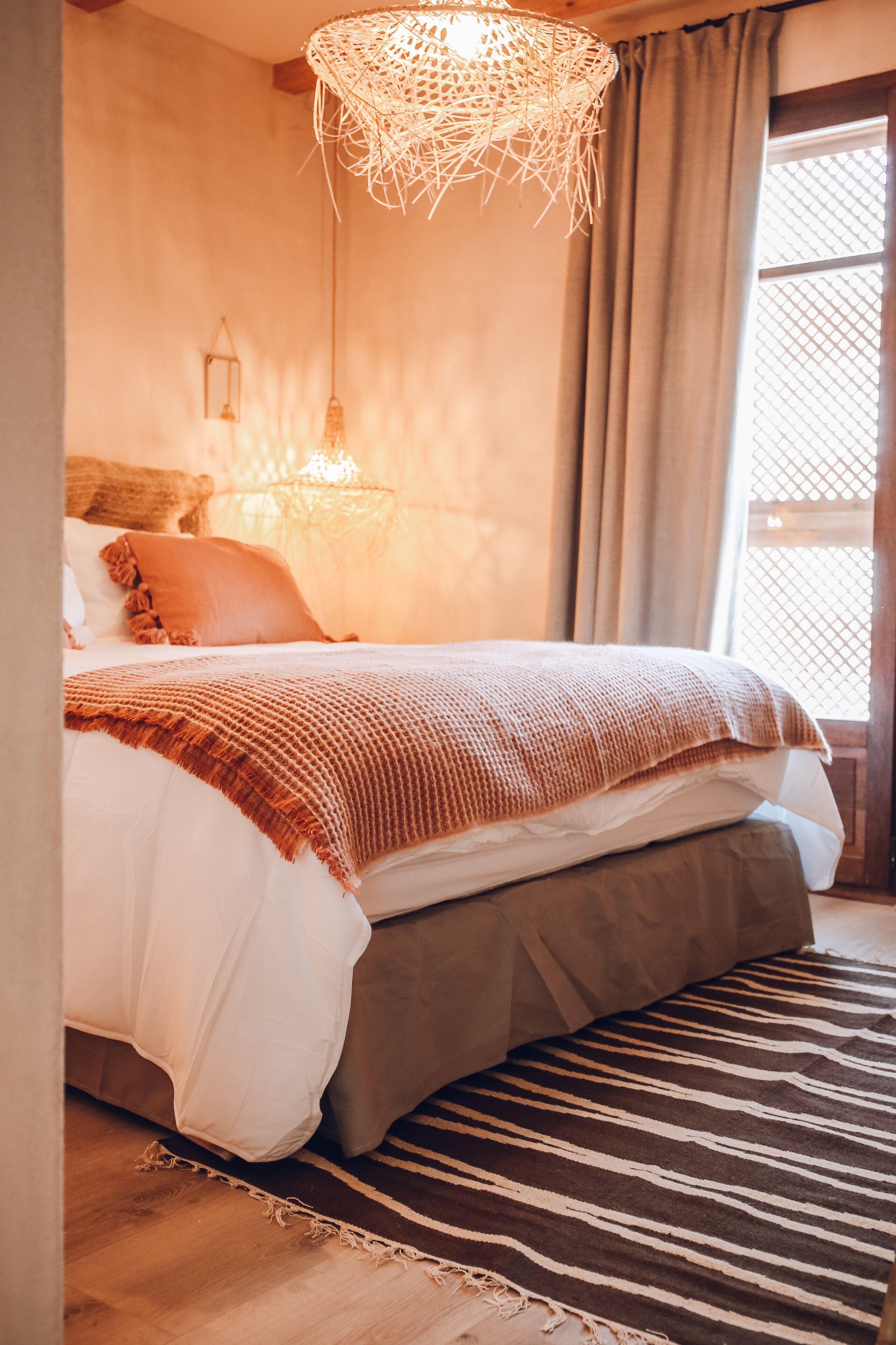 A neatly made bed with a striped rug, a woven light fixture, and a window with curtains.