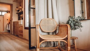 Una silla de mimbre con respaldo y asiento tejido, ubicada cerca de una ventana con cortinas traslúcidas, un piso de madera y un espejo en la pared.