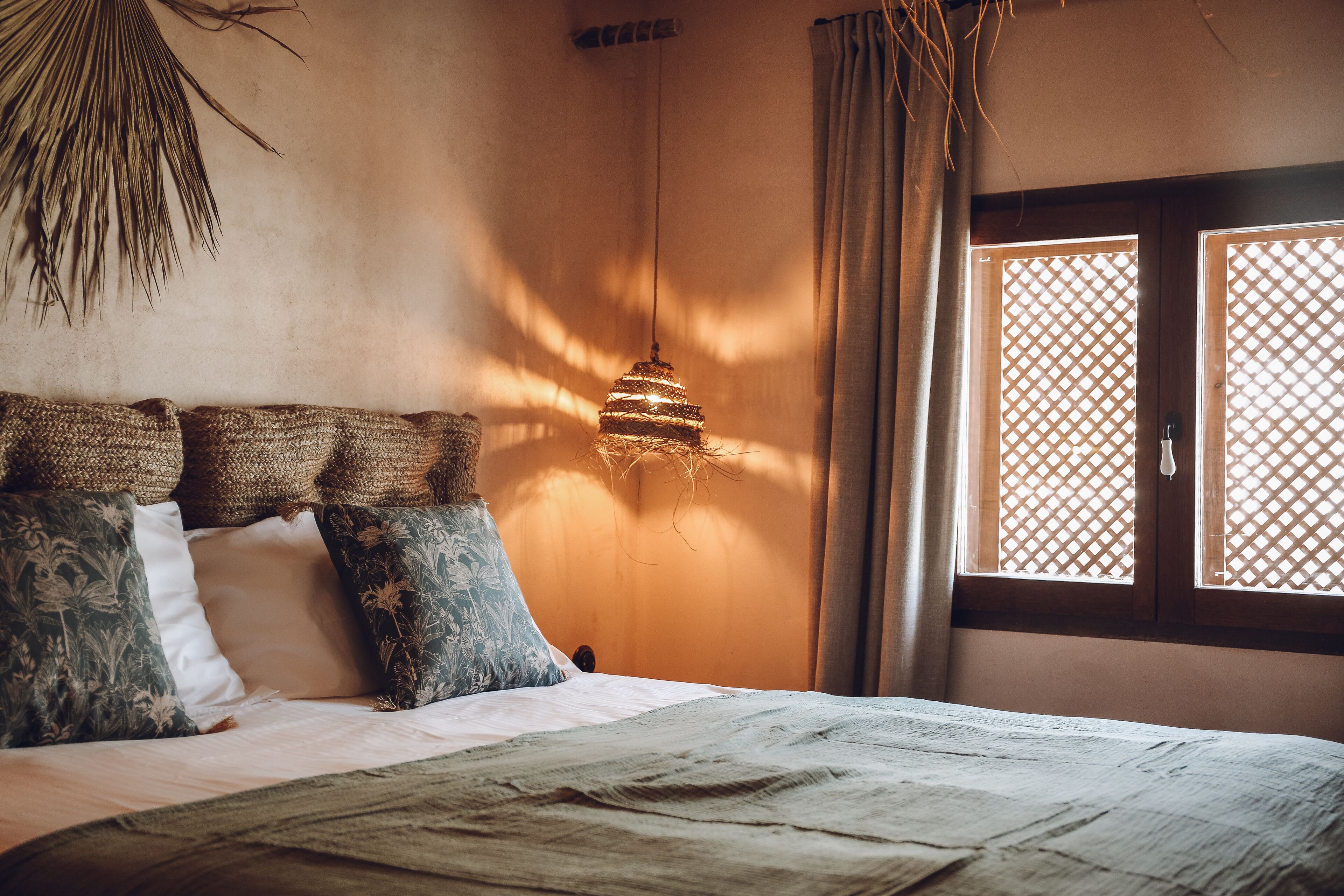 A bedroom with a bed, a hanging light fixture, a window with a lattice screen, and a wall-mounted fan.