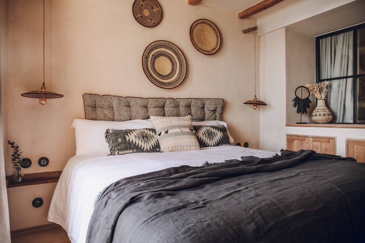 Ein Schlafzimmer mit einem gepolsterten Kopfteil, einem Bett mit weißer Bettwäsche, einer dunkelgrauen Tagesdecke und einem Nachttisch mit einer Vase getrockneter Blumen.