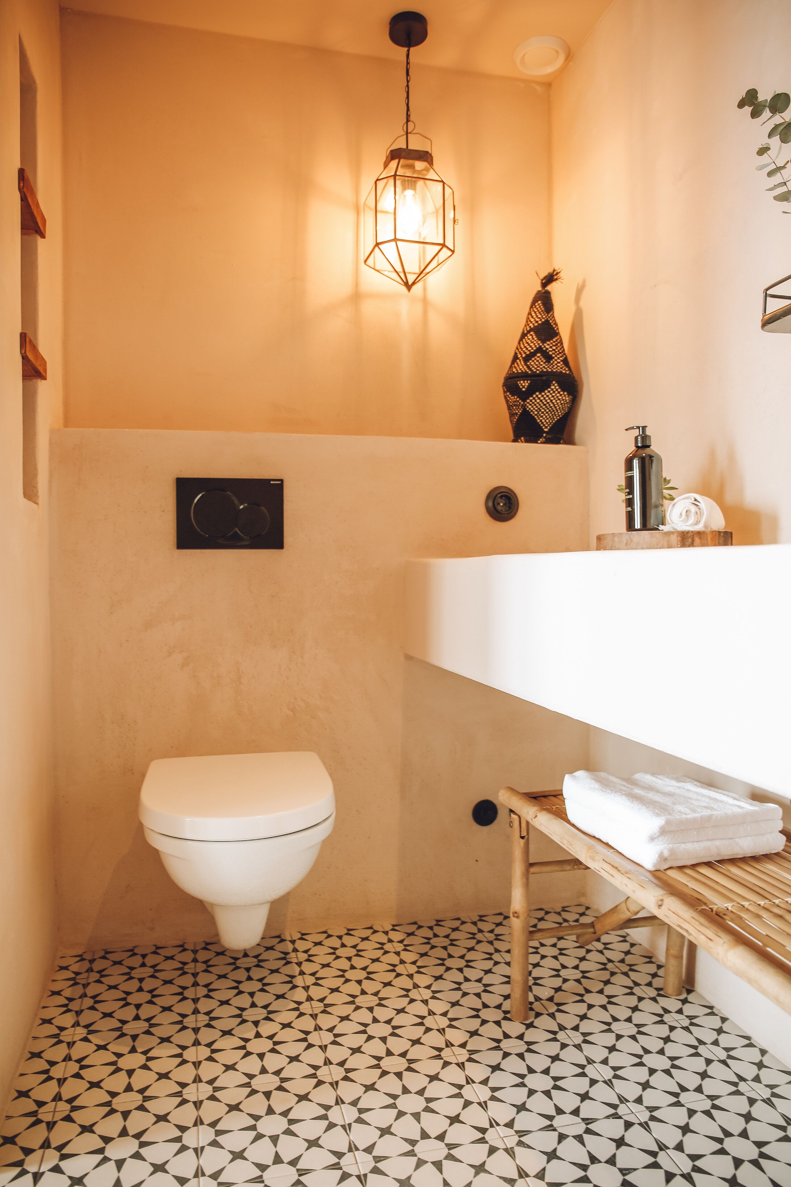 A compact bathroom with a white toilet, a wooden bench, a white sink, and a geometric patterned floor.