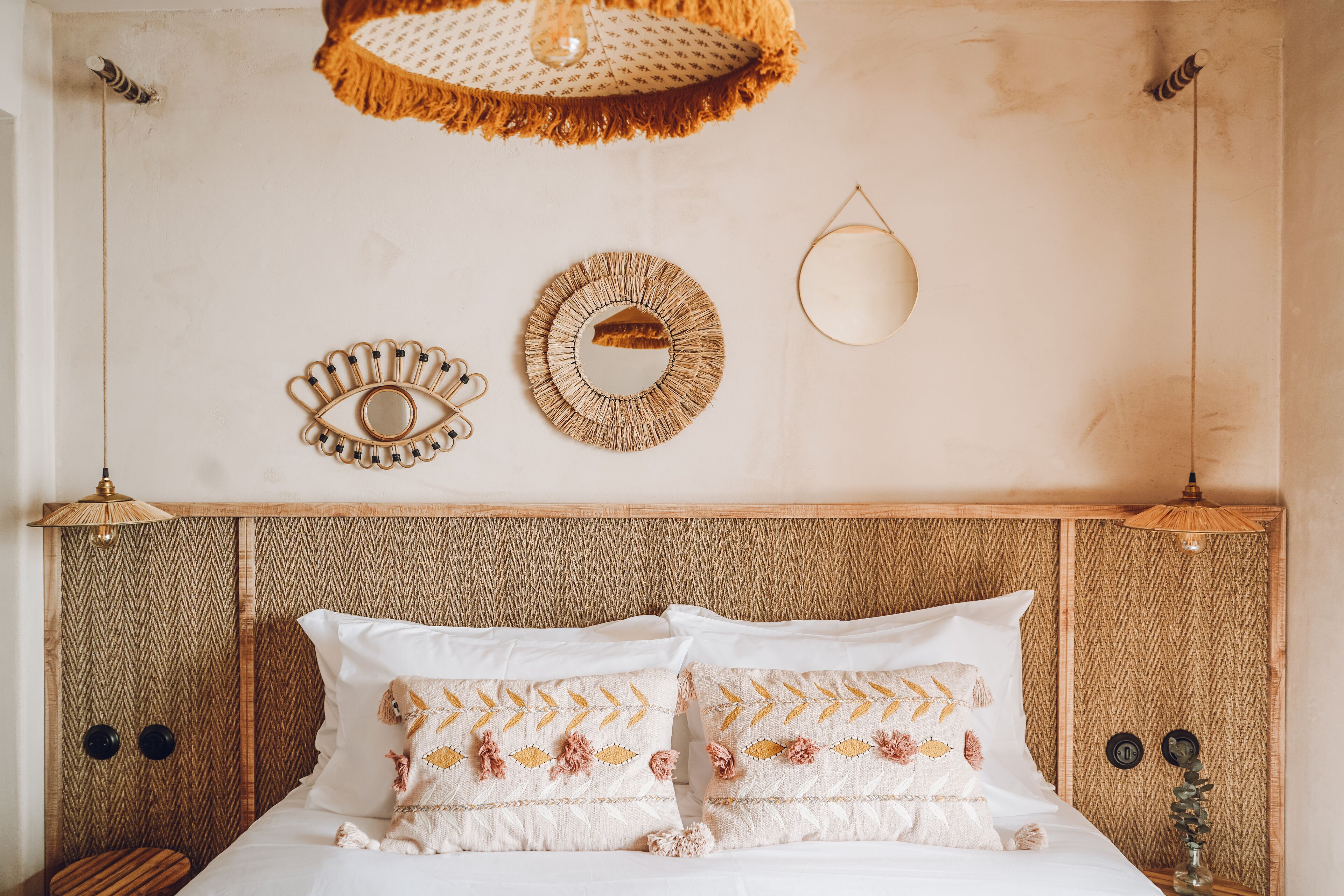 A bed with white linens and decorative pillows, a woven headboard, and wall-mounted mirrors and light fixtures.