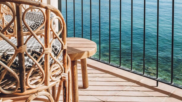 Una terraza de madera con una silla de mimbre y una mesa pequeña, con vista a un mar tranquilo y un litoral rocoso.