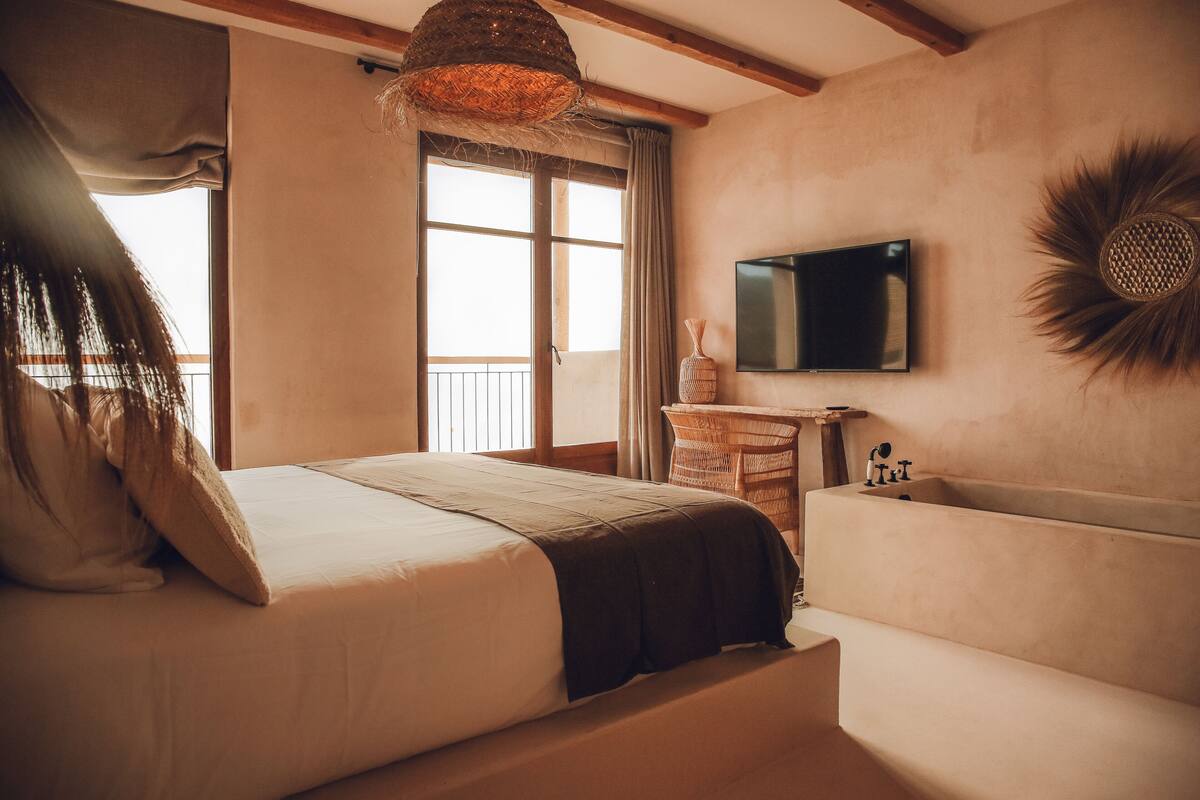 Ein Schlafzimmer mit einem Bett, einem an der Wand befestigten Fernseher, einem Fenster mit Vorhängen und einer Holzbalkendecke.