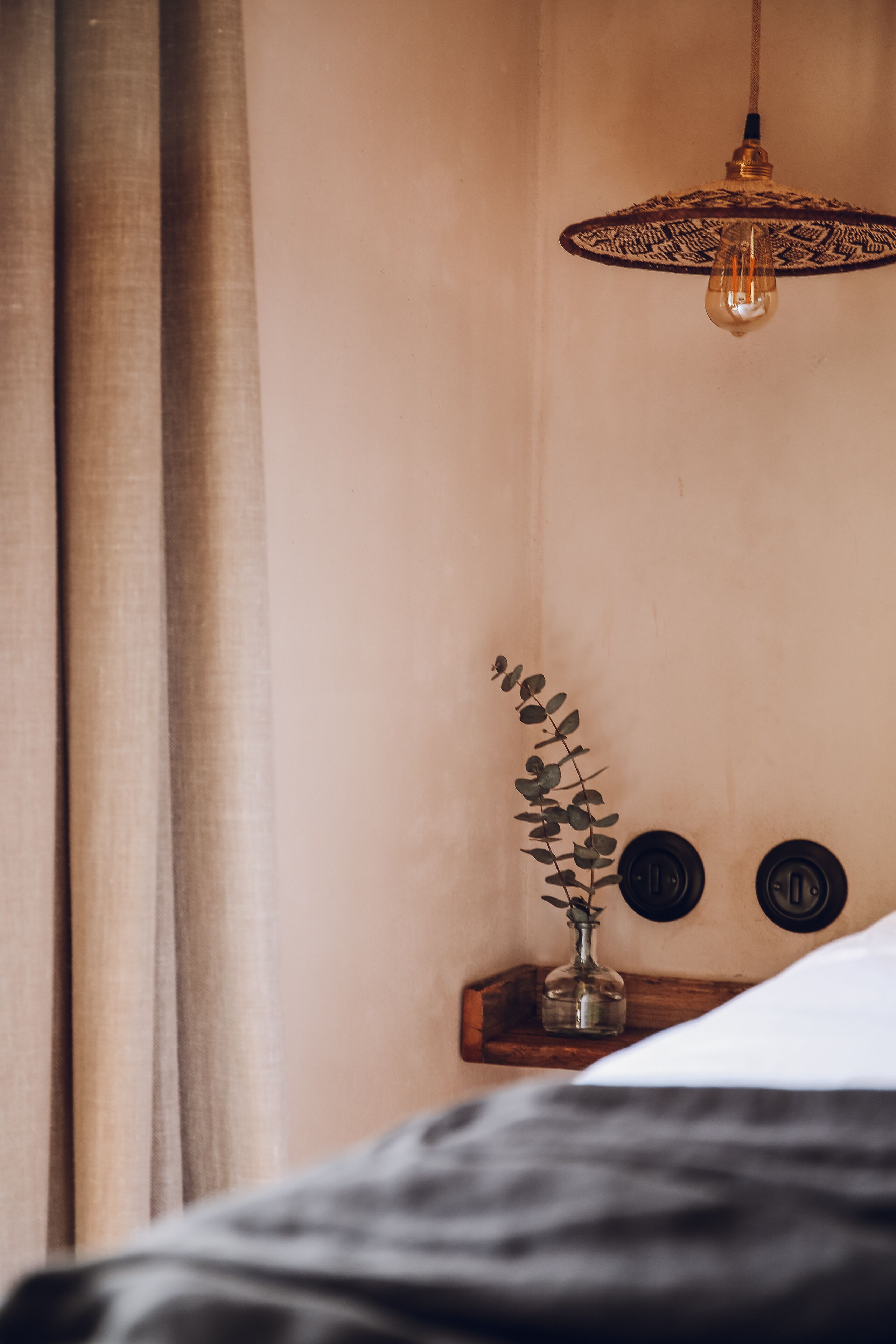 A bedroom with a bed, a hanging light fixture, a small wooden shelf with a plant, and two round wall decorations.