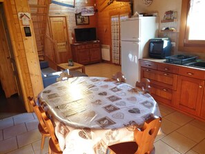 Une chambre confortable, aménagée dans le style d’un chalet, avec une table à manger en bois, des chaises, un petit coin cuisine équipé d’un réfrigérateur, une télévision et une fenêtre avec des rideaux.