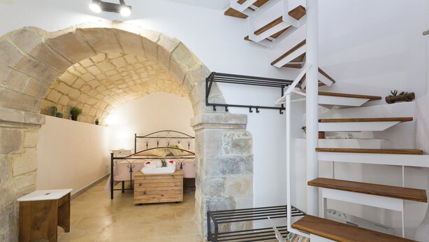 A stone-walled room with a bed, a wooden bench, and a metal drying rack.
