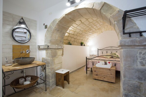 A rustic bathroom with a stone archway, a wooden vanity with a bowl sink, a small stool, and a bed with a metal frame.