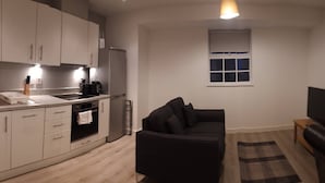 A modern kitchen with white cabinets, a black countertop, and a built-in oven. There is a black sofa and a small wooden table with a TV in the living area. - Riverside 4 (Maidstone)