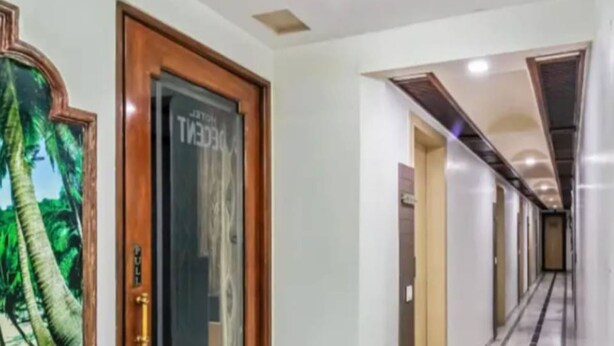 A hotel corridor with a wooden door, a framed picture of a palm tree, and a tiled floor. - Hotel Decent (Surat)
